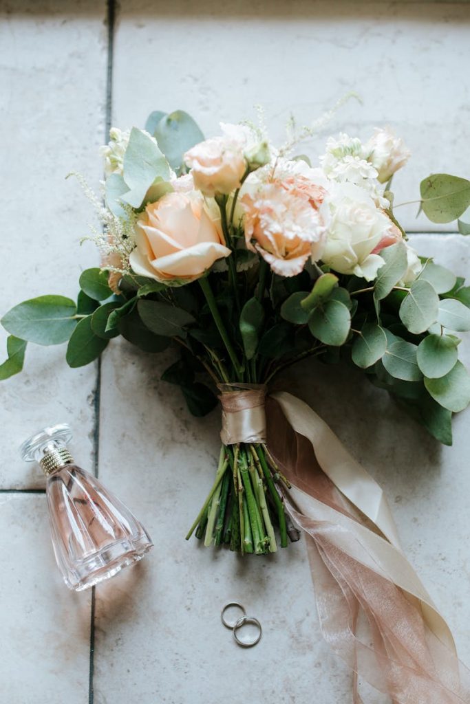 Beautiful wedding bouquet with roses, rings, and perfume bottle arranged on a tiled floor.